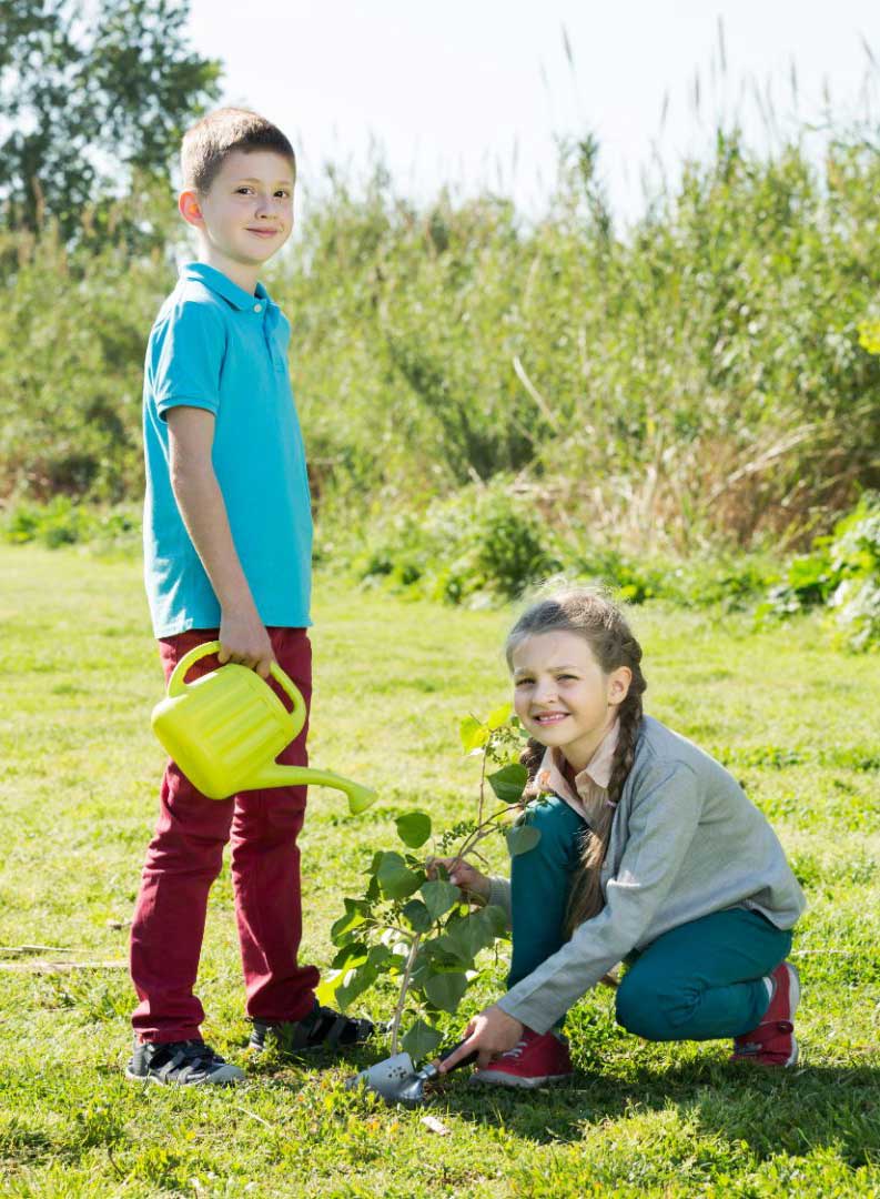 girl kneeling on ground next to a tree by a boy standing up and holding a watering can