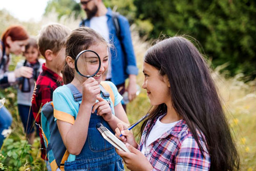 photo of kids in a field with one girl looking through a magnifiying glass at another girl looking towards her