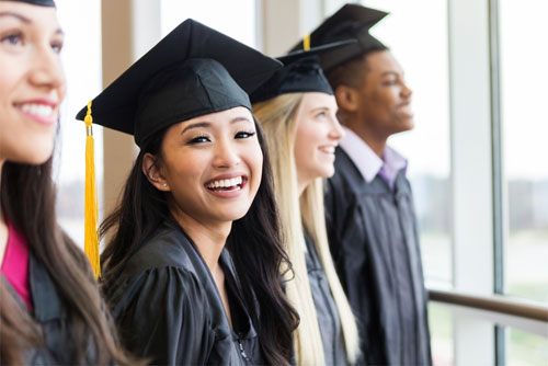 photo of graduates standing in a row with black gowns and mortarboards with one Asian girl turned toward the camera and smiling