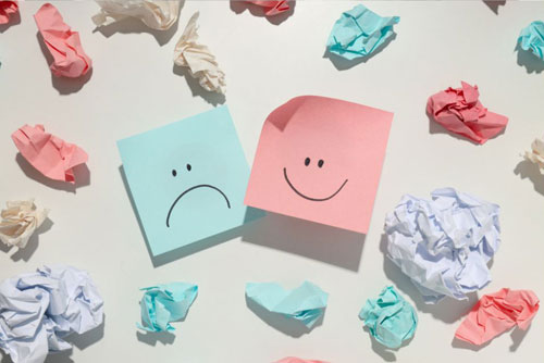 photo of a pink Post It note with a smiley face next to a light green Post It note with a frown face surrounded by crumbled up pieces of paper